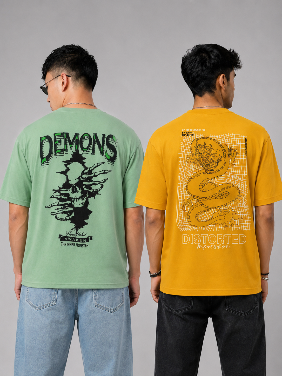 Two men shown from the back wearing graphic t-shirts. The man on the left wears a light green t-shirt with a black 'DEMONS' skull graphic, and the man on the right wears a mustard yellow t-shirt with a beige snake graphic.