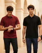 Two men walking side by side on a cobblestone street in front of a warm, lit storefront. The man on the left wears a burgundy polo shirt and dark trousers, while the man on the right wears a black polo shirt and navy trousers.