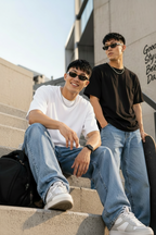 Two male models wearing oversized sunglasses, casual tees, and jeans pose on outdoor stone steps with a black bag beside them. The image is a promotional graphic for a pack of two tees from Mavincub, featuring text overlays and product details.