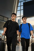 Two young men walking with rolling suitcases in an airport terminal, wearing casual streetwear. The man on the left wears a black t-shirt, black pants, and a black backpack.